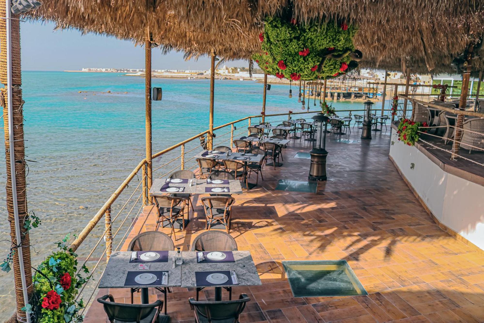 泻湖畔豪华spa度假村 麦纳麦 外观 照片 The photo shows a seaside restaurant with a spacious outdoor dining area. It features several tables set with plates and utensils, surrounded by lush greenery and decorative plants. The flooring is made of brown tiles, and there are thatched roofs pr
