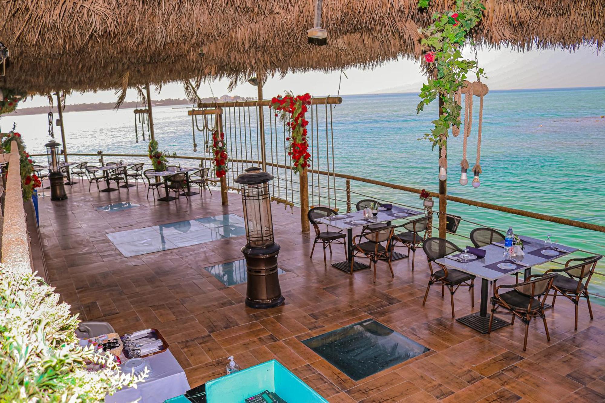 泻湖畔豪华spa度假村 麦纳麦 外观 照片 The photo depicts a coastal restaurant or dining area with an open-air design. It features a wooden floor and tables set for dining, surrounded by chairs. The space is partially covered by a thatched roof, providing shade while allowing for a view of