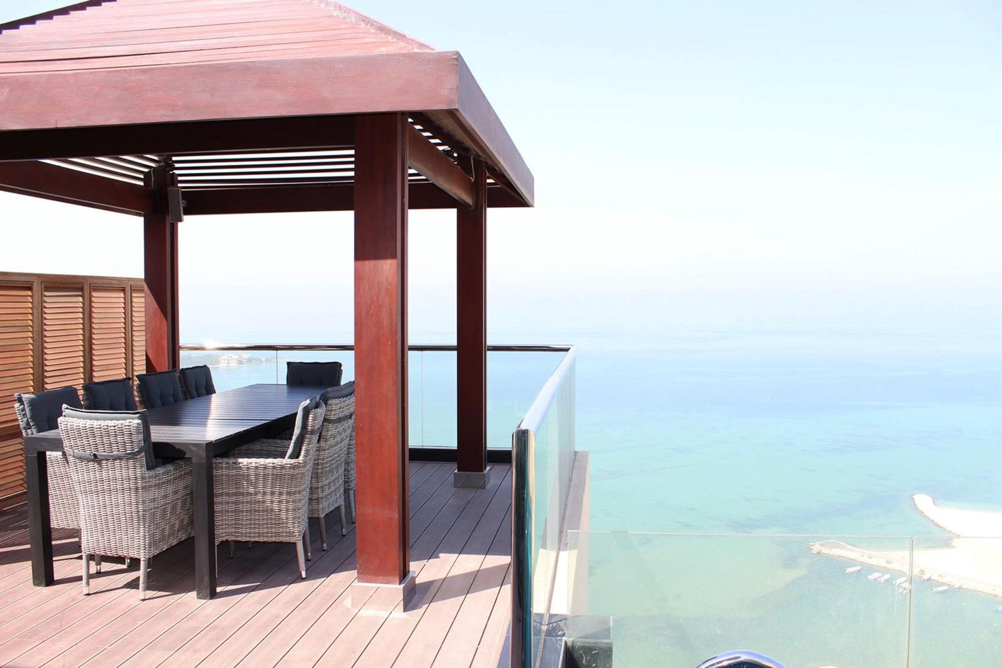 泻湖畔豪华spa度假村 麦纳麦 外观 照片 The photo showcases a stylish outdoor patio or terrace area. It features a wooden structure with a sloped roof, providing a shaded space. There is a large table made of dark wood, surrounded by several wicker chairs. The patio is elevated and has gla