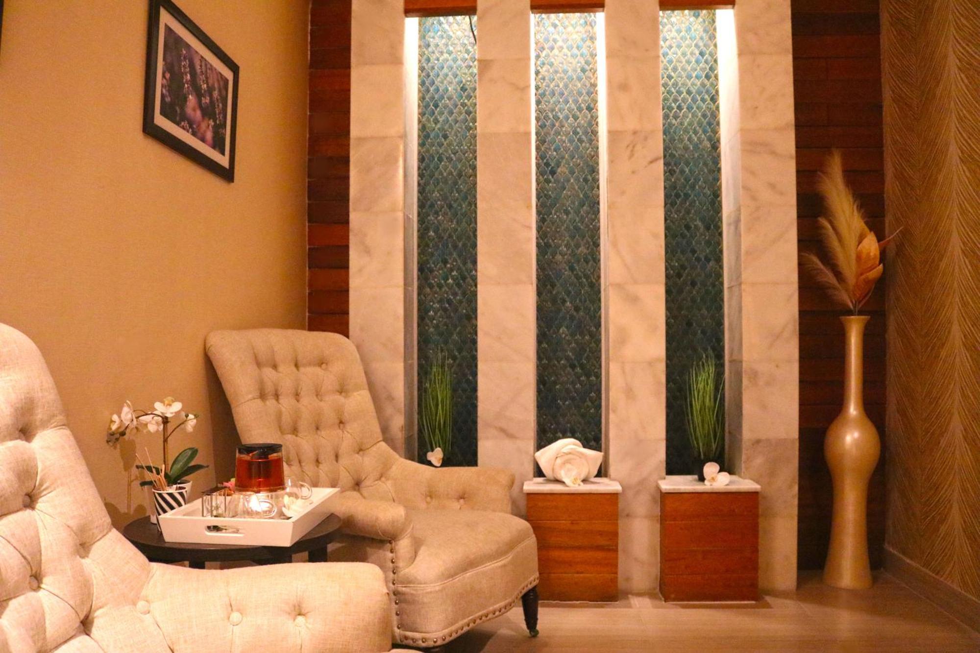 泻湖畔豪华spa度假村 麦纳麦 外观 照片 The image shows a relaxing interior space, likely a waiting area or lounge. There are two plush, beige armchairs positioned near a small table, which holds a glass pitcher and a cup. The background features tall, elegant windows with decorative blue 