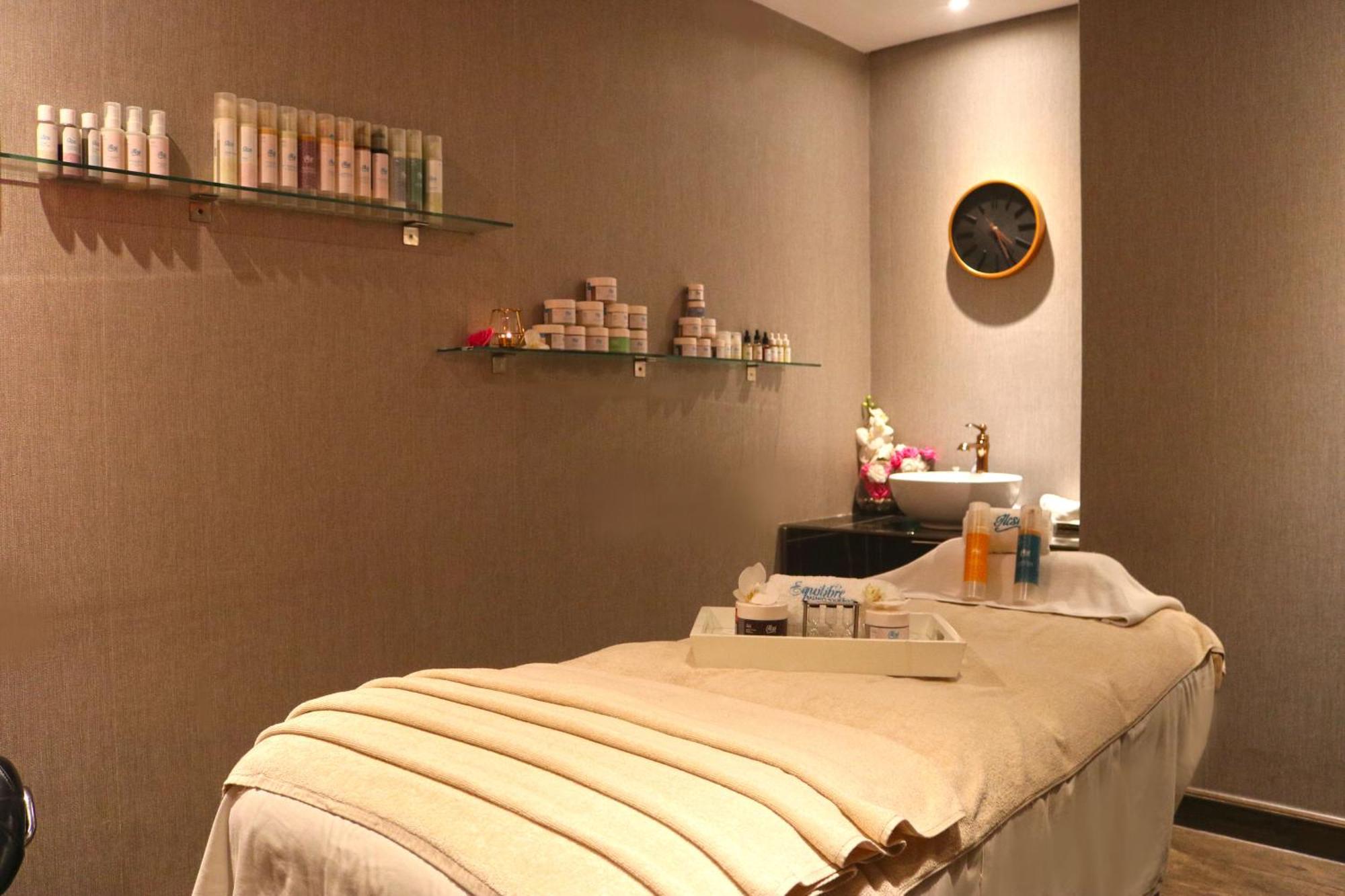 泻湖畔豪华spa度假村 麦纳麦 外观 照片 The photo shows a spa treatment room. There is a massage table draped with a light-colored blanket, creating a relaxing atmosphere. On the wall, there are shelves displaying various skincare products in bottles and jars. A clock is mounted on the wal
