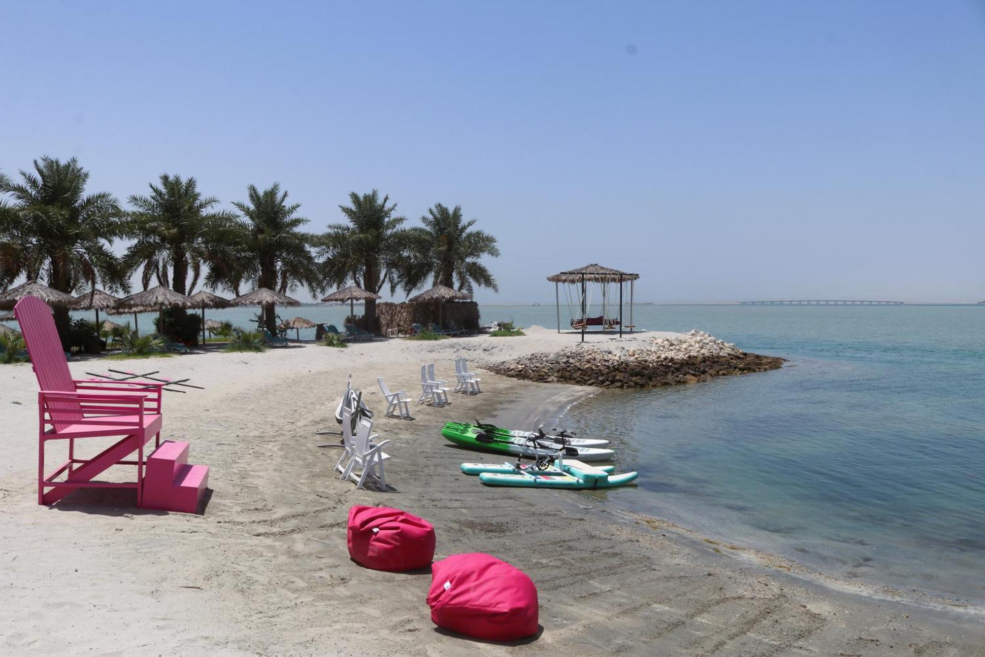 泻湖畔豪华spa度假村 麦纳麦 外观 照片 The photo depicts a serene beach scene with clear blue water and a sunny sky. In the foreground, there are colorful pink bean bags and a large pink chair on the sand. A few white chairs and loungers are positioned near the water's edge. To the side, 