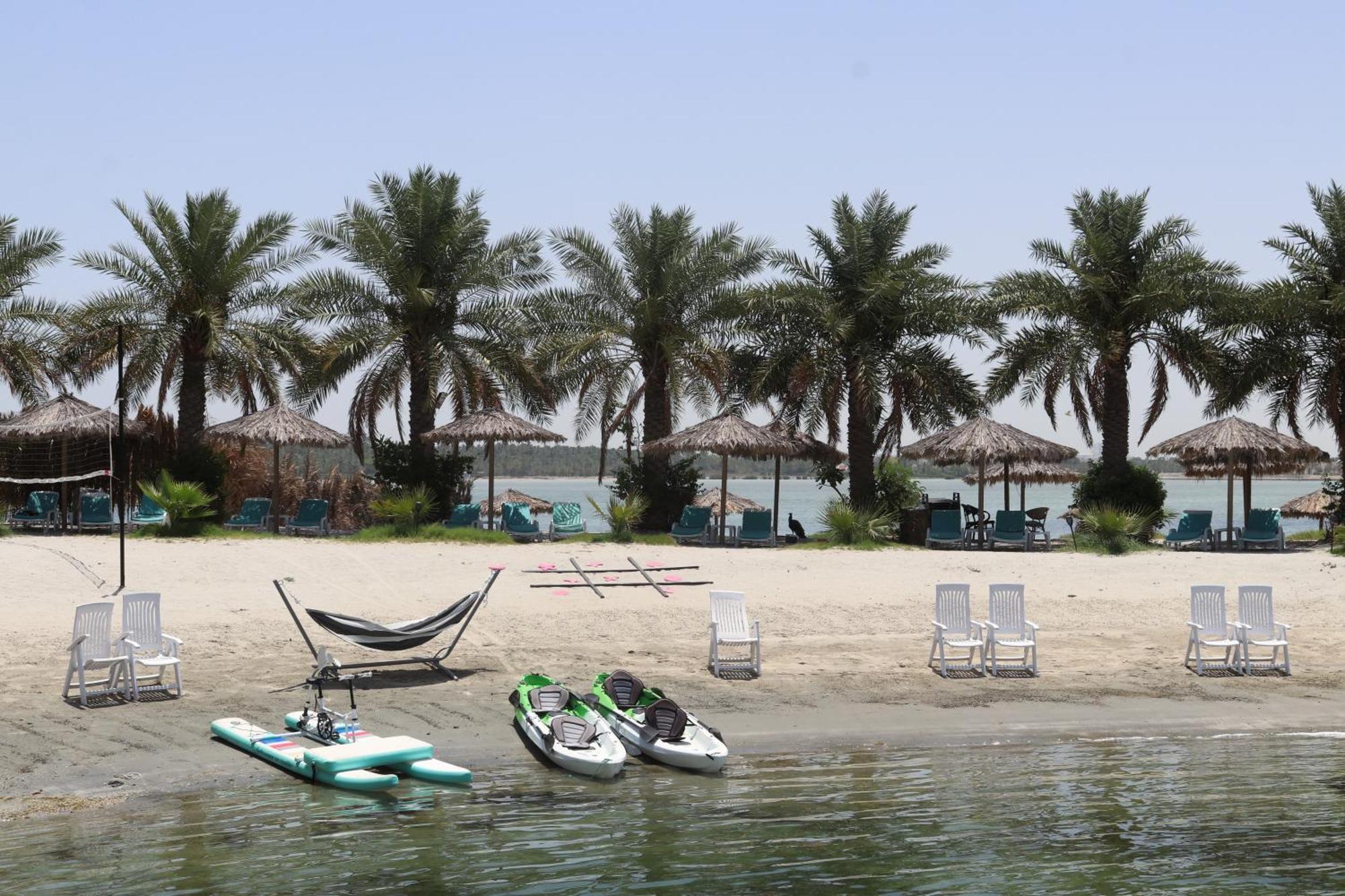 泻湖畔豪华spa度假村 麦纳麦 外观 照片 The photo depicts a serene beach scene with several palm trees lining the shore. There are thatched umbrellas providing shade, and a few lounge chairs are positioned on the sand. A hammock is set up between two supports, inviting relaxation. In the f