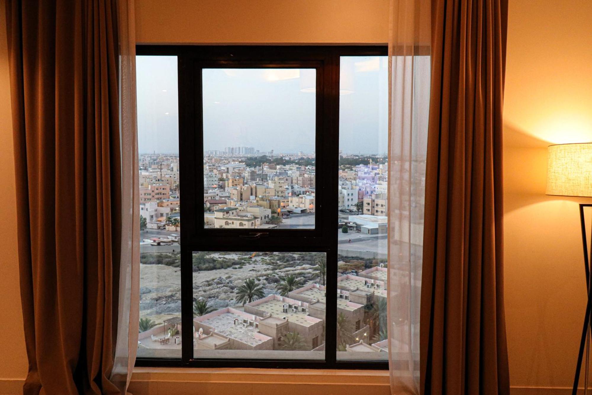 泻湖畔豪华spa度假村 麦纳麦 外观 照片 The photo shows a window with a black frame and sheer curtains. Through the window, there is a view of a cityscape, displaying a densely populated area with various buildings and rooftops. The sky appears slightly overcast, suggesting a muted lightin