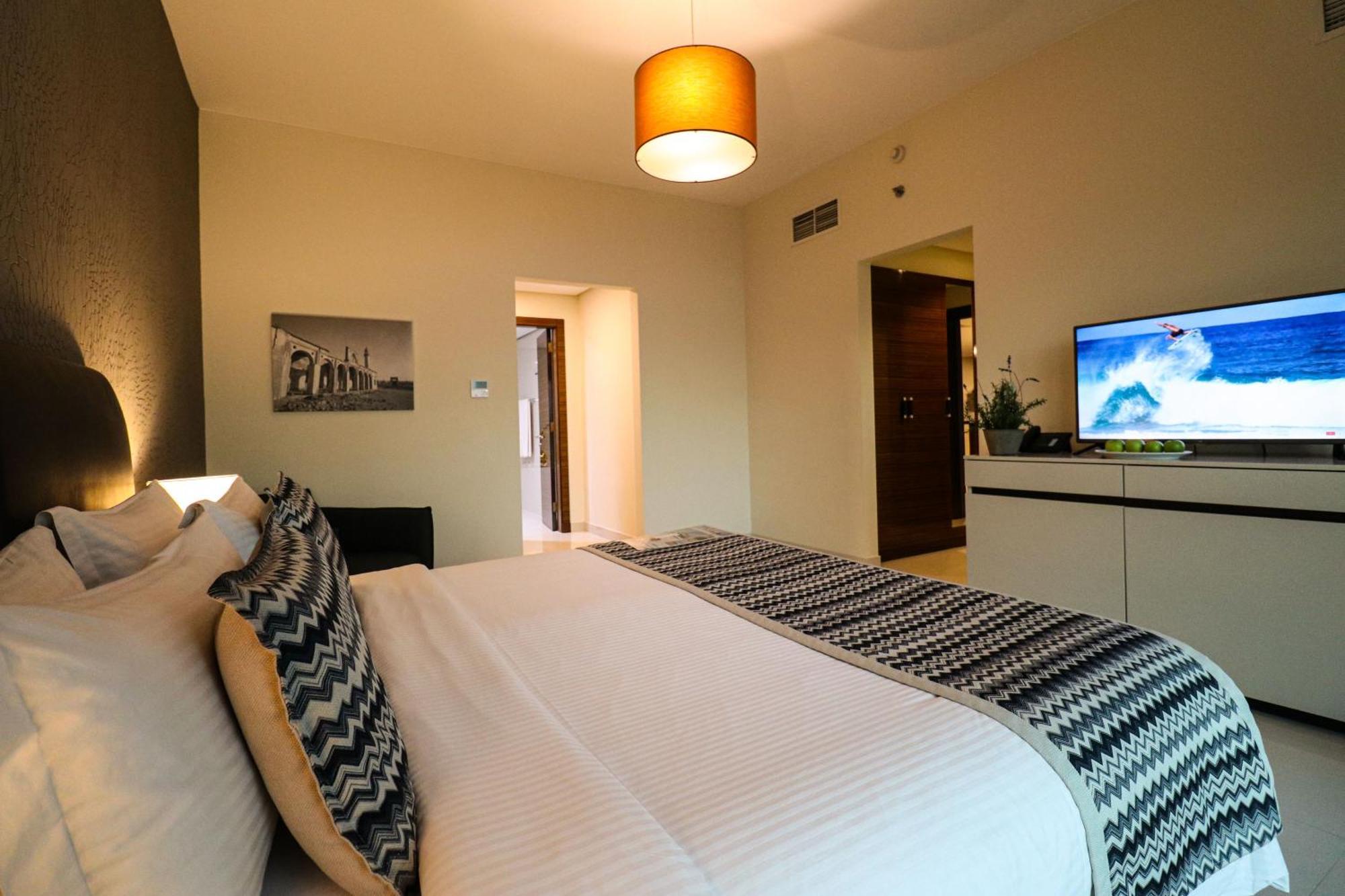 泻湖畔豪华spa度假村 麦纳麦 外观 照片 The photo shows a well-decorated hotel room. There is a large bed with white linens and decorative pillows featuring a zigzag pattern. On the wall beside the bed is a piece of artwork, and there is a modern television on a white furniture unit opposi
