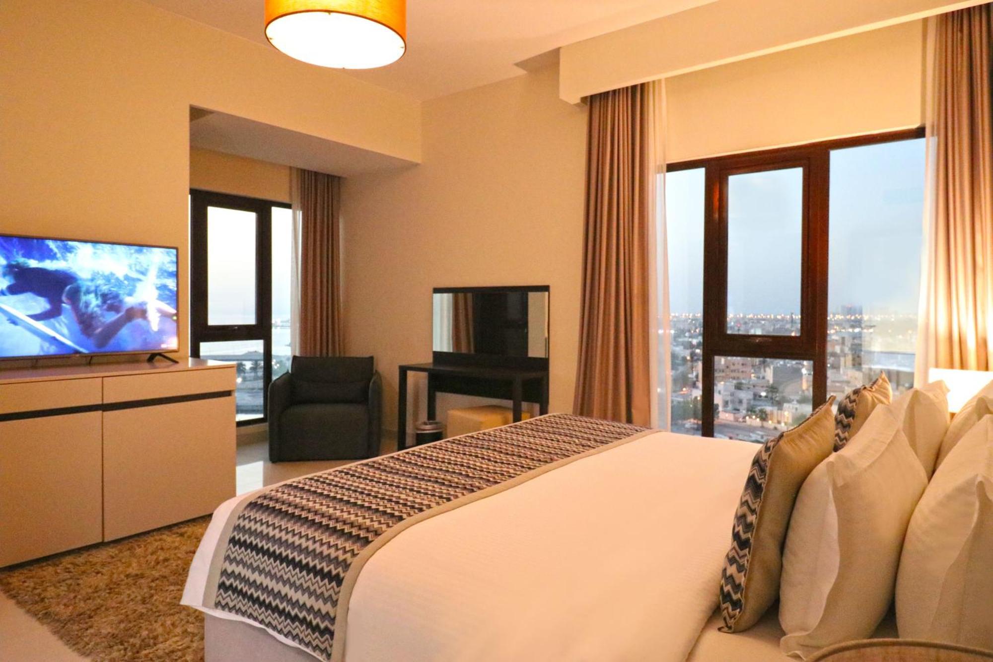 泻湖畔豪华spa度假村 麦纳麦 外观 照片 The photo shows a modern hotel room or suite. It features a neatly made bed with decorative pillows in the foreground. To the left, there is a TV on a cabinet, and further back, a comfortable chair is situated in front of a large window that likely o