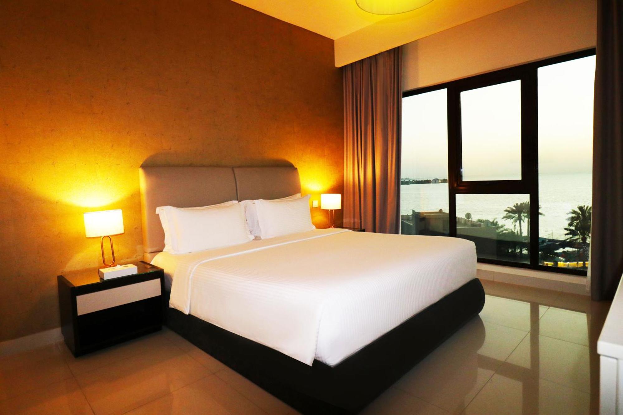泻湖畔豪华spa度假村 麦纳麦 外观 照片 The photo shows a modern hotel bedroom with a large, comfortable bed dressed in white linens. The room features warm lighting, including bedside lamps, creating a cozy atmosphere. There are large windows with a view of the outdoors, possibly overlook